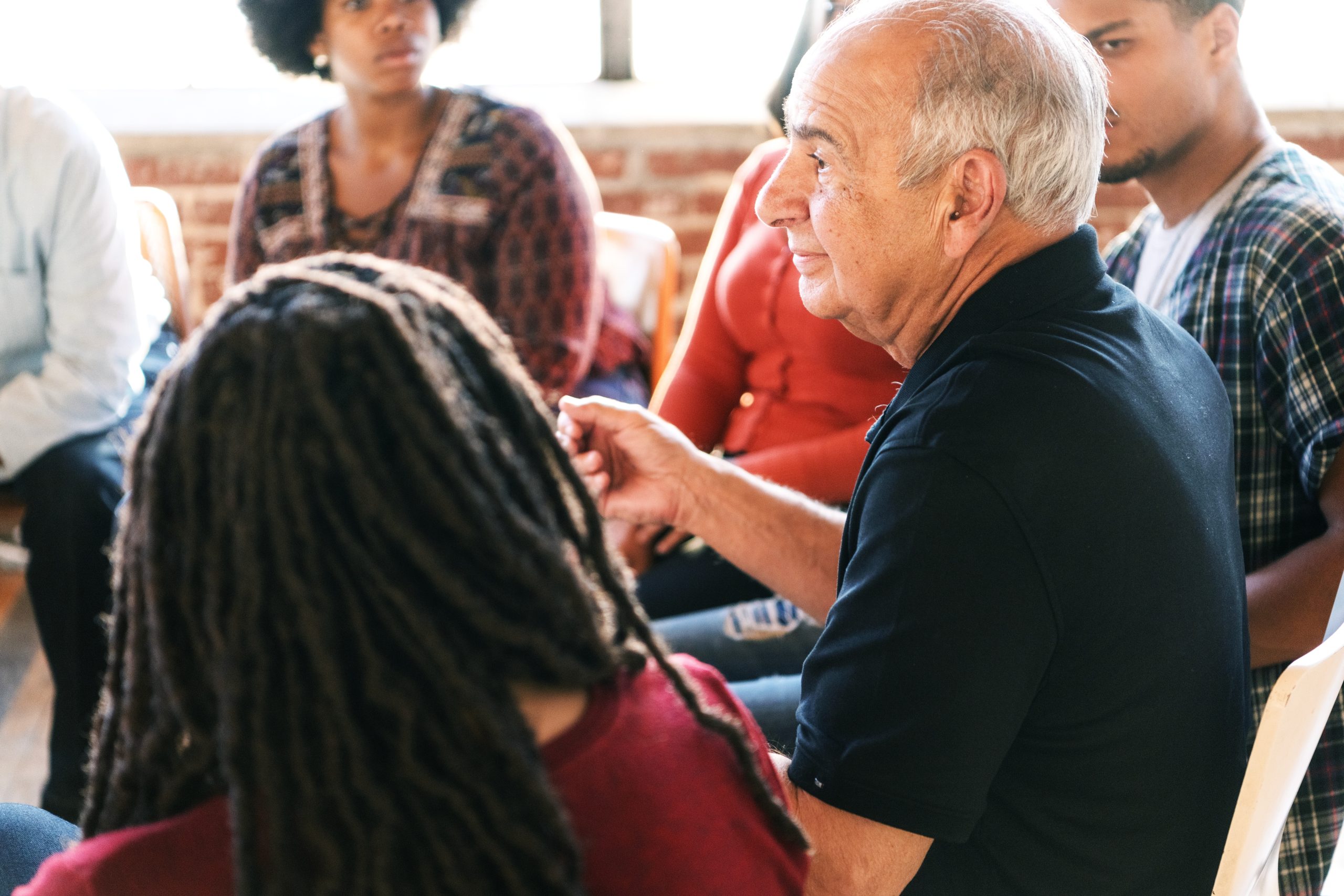 A diverse group of people in a discussion circle, including an older man speaking. The group listens attentively, fostering communication.
